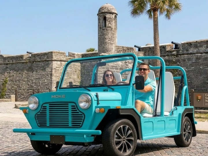 Two people in an open turquoise car near an old stone fortress.