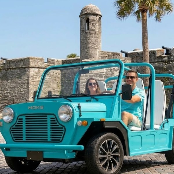 Two people in an open turquoise car near an old stone fortress.