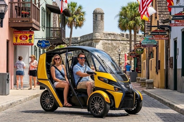 Couple rides a small, yellow, three-wheeled vehicle on a historic street with shops and flags.
