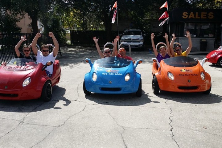 People in small colorful cars raising their hands at a sales location.