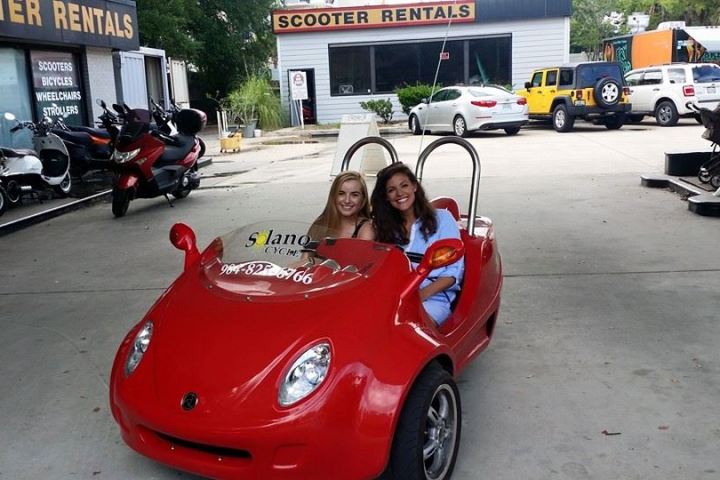 Two people smiling in a red open-top car at a scooter rental shop.