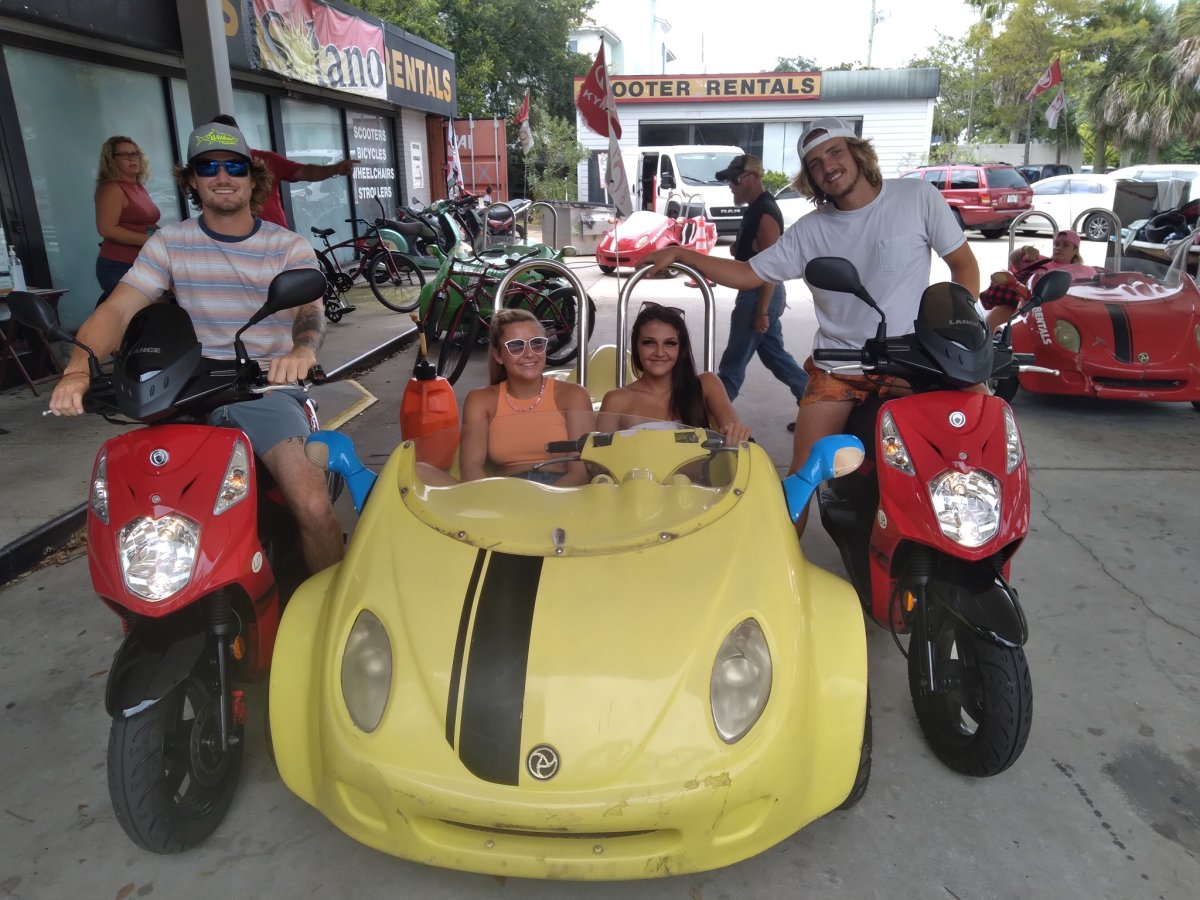 Four people posing with two scooters and a yellow three-wheeler in front of a rental shop.