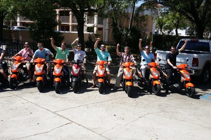 Group of nine people on orange scooters, raising arms, parked in a sunny lot.