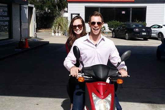 Two people on a red scooter in front of a scooter rentals shop.